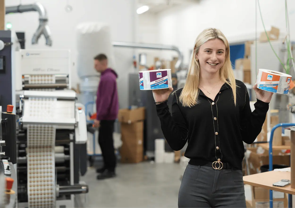 Mitarbeiterin steht in der Produktion und hält zwei Etikettenrollen hoch.